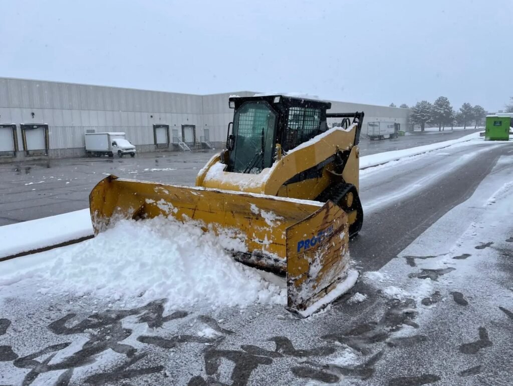 Yellow compact track loader with a snow pusher clearing a commercial parking lot beside a warehouse